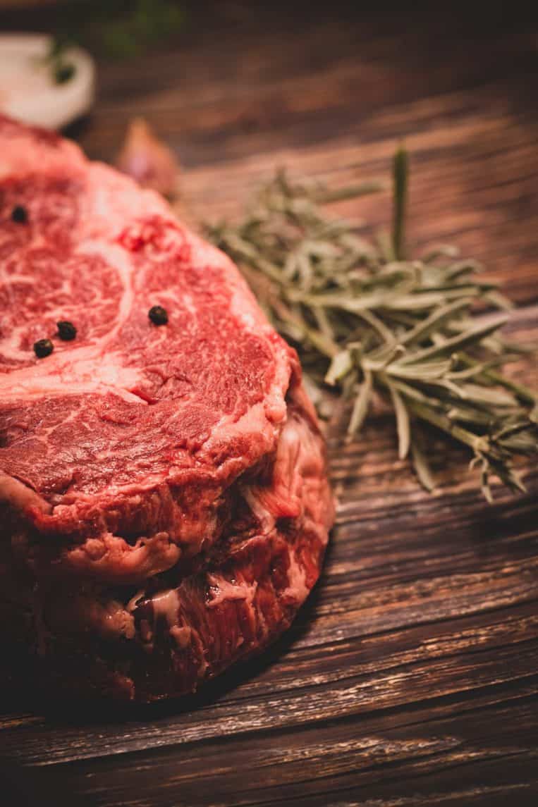 Uncooked steak with rosemary and peppercorns on wood.