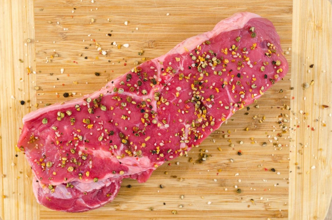 A raw steak seasoned with crushed peppercorns and coarse salt on a wooden cutting board with loose seasoning scattered around