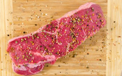 A raw steak seasoned with crushed peppercorns and coarse salt on a wooden cutting board with loose seasoning scattered around