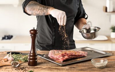 a male cook sprinkles spices on a piece of meat resting on a tray