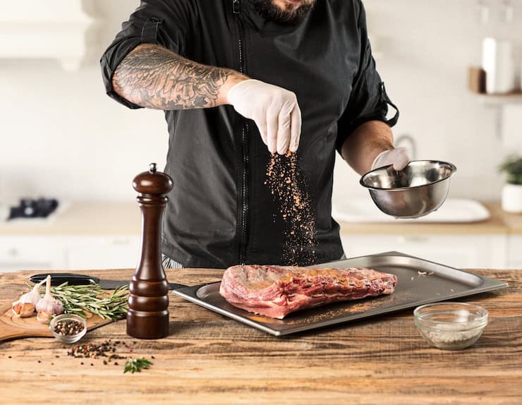 a male cook sprinkles spices on a piece of meat resting on a tray